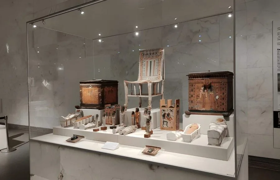 A glass display case in the National Museum of Egyptian Civilization (NMEC) filled with various ancient Egyptian artifacts including wooden chairs, miniature coffins, and small statues.