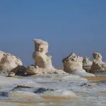 A group of massive, wind-eroded, white chalk rock formations standing like sculptures in the sandy, desolate landscape of the White Desert National Park in Egypt under a bright blue sky.