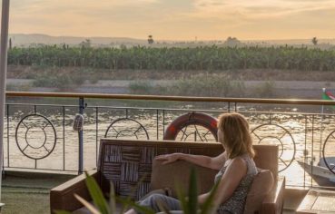 A Woman Contemplating The View From A Nile River Boat, sitting on the deck enjoying the sunset and the view of the lush, green riverbanks.