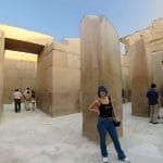 A modern Woman poses between the huge, polished stone pillars of the ancient Valley Temple of Khafre in Giza, Egypt, with other tourists in the background.