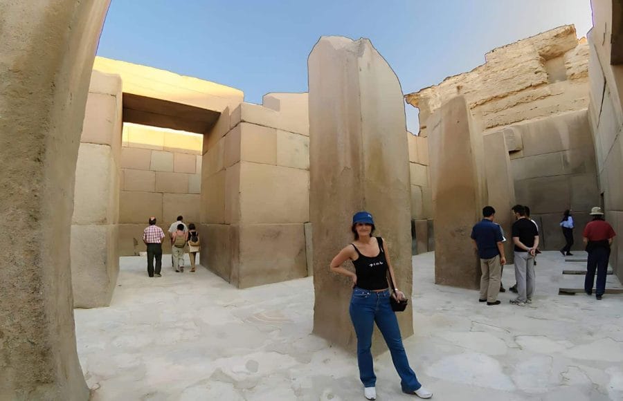 A modern Woman poses between the huge, polished stone pillars of the ancient Valley Temple of Khafre in Giza, Egypt, with other tourists in the background.
