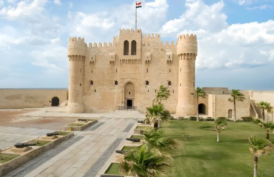 The imposing stone exterior of the Citadel of Qaitbay Amphitheatre in Alexandria, showing its medieval fortress walls, battlements, and central courtyard with cannons.