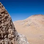 A close-up view of large, jagged crystals protruding from the earth at Crystal Mountain in the White Desert National Park, Libyan Desert, set against a backdrop of sandy hills and a blue sky.