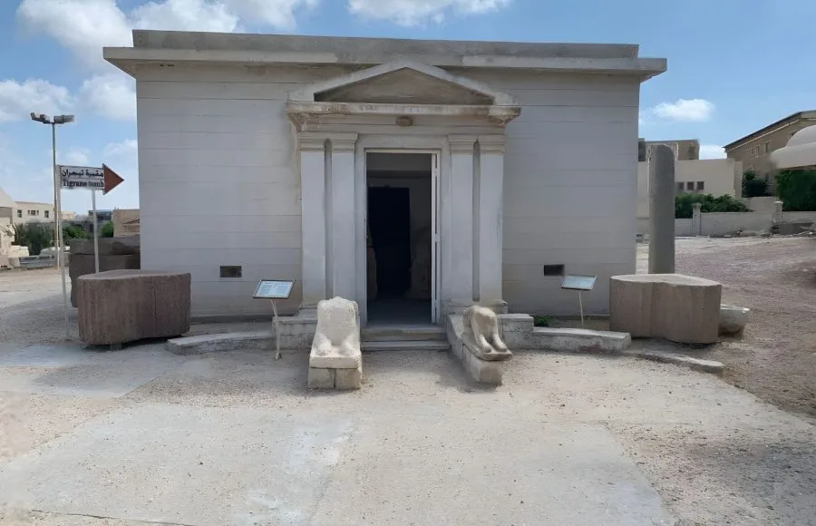 The modern, light-colored facade of the entrance to the Tigran Tomb at Alexandria, located near the site of Kom el-Shoqafa, with an open doorway and stone fragments in the foreground.