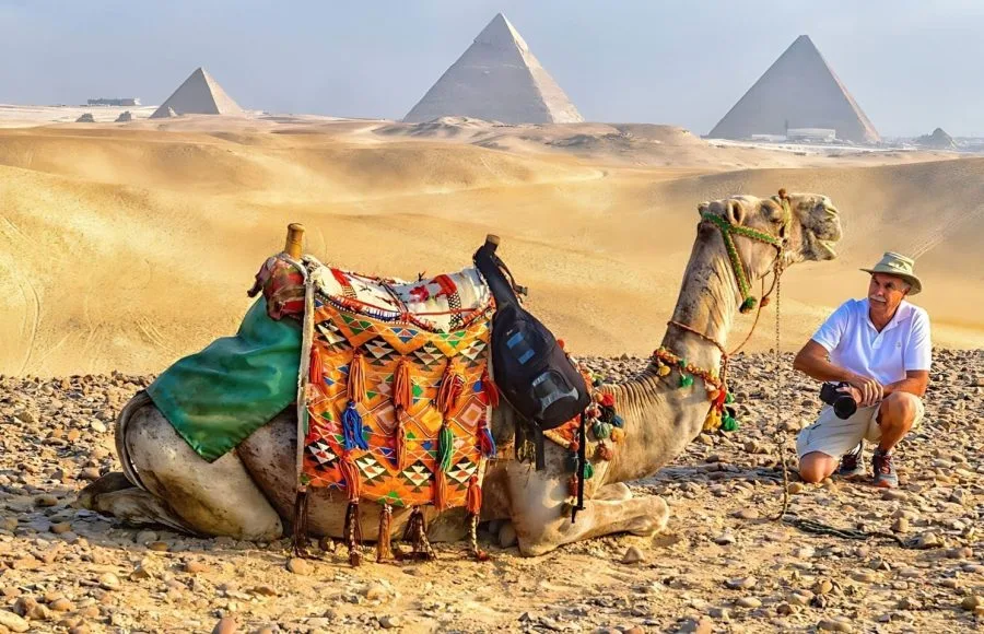 A tourist kneeling next to a saddled camel, with the backdrop of the Pyramids of Giza during a Camel Ride Through the Desert, highlighting the classic Egyptian desert experience.