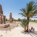 The colossal, weathered stone Colossi of Memnon luxor, showing the two seated statues of Amenhotep III with tourists and a date palm in the foreground.