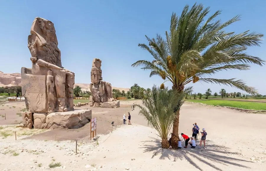 The colossal, weathered stone Colossi of Memnon luxor, showing the two seated statues of Amenhotep III with tourists and a date palm in the foreground.