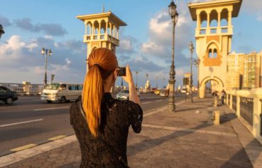 A Female Tourist With photographing Stanley Bridge Rear View Alexandria Egypt, seen from behind on the pedestrian walkway, capturing the ornate towers of the bridge at sunset.
