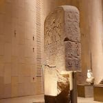A large, ancient Egyptian stela covered in hieroglyphs and relief carvings, displayed under dramatic lighting inside the modern Gem Grand Egyptian Museum New Giza Museum (GEM).