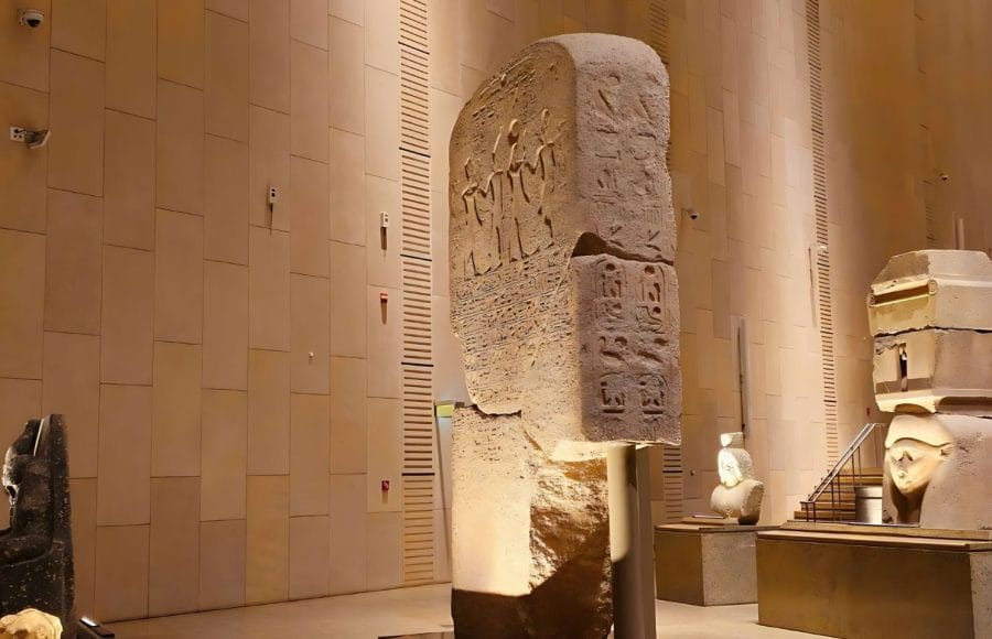 A large, ancient Egyptian stela covered in hieroglyphs and relief carvings, displayed under dramatic lighting inside the modern Gem Grand Egyptian Museum New Giza Museum (GEM).