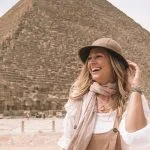 A beautiful, smiling Lady Wearing Hat stands in front of the colossal stone blocks of one of the Giza Pyramids.