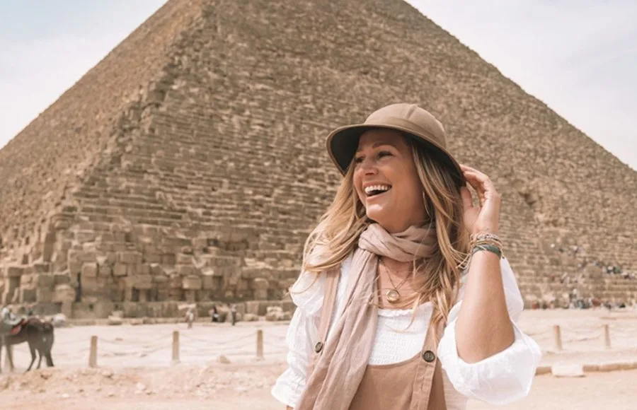 A beautiful, smiling Lady Wearing Hat stands in front of the colossal stone blocks of one of the Giza Pyramids.