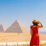 A Girl in a flowing Red dress and sunhat stands with her back to the camera, admiring the majestic Giza Pyramids in the desert under a clear blue sky.