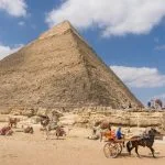 The Great Pyramid of Giza under a blue sky, with a horse-drawn cart and resting camels.