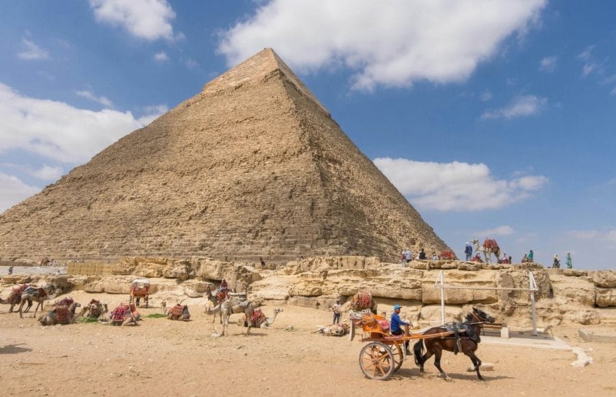 The Great Pyramid of Giza under a blue sky, with a horse-drawn cart and resting camels.