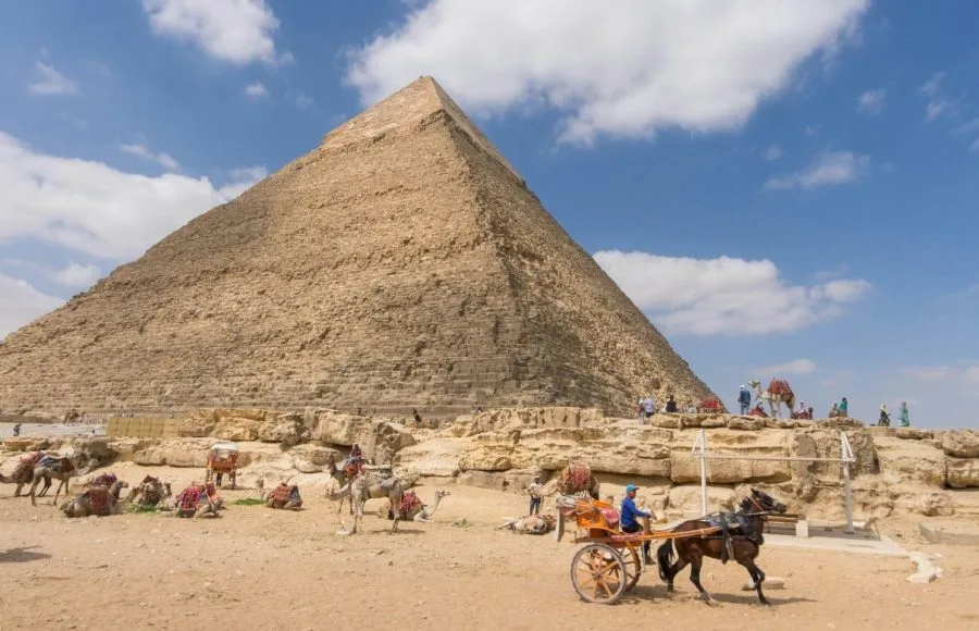 The Great Pyramid of Giza under a blue sky, with a horse-drawn cart and resting camels.