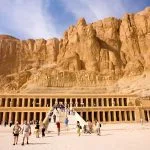 A large group of tourists at the Temple of Hatshepsut at Deir el-Bahari, showing its terraced structure against the backdrop of towering cliffs.