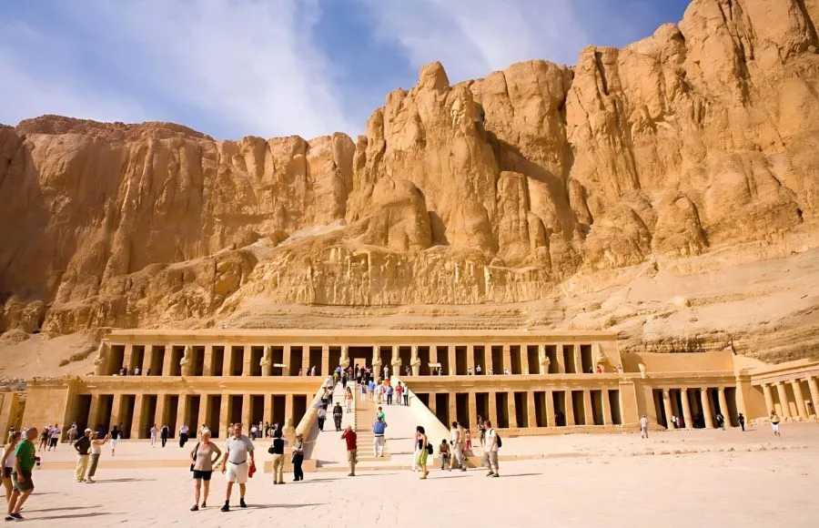 A large group of tourists at the Temple of Hatshepsut at Deir el-Bahari, showing its terraced structure against the backdrop of towering cliffs.