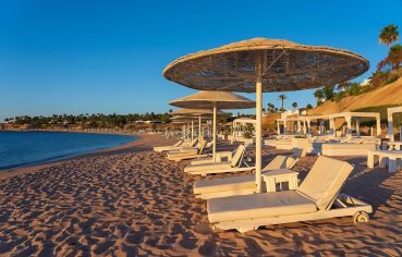 A beautiful, wide Hurghada Sand Beach at sunrise or sunset, featuring a row of white sun loungers and straw umbrellas.
