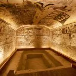 A view Inside Valley of the Kings showing a burial chamber with brightly painted and well-preserved scenes and hieroglyphs on the walls and ceiling, including a large rectangular sarcophagus pit.