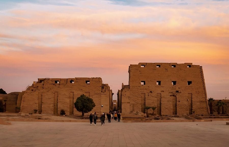 The massive stone entrance pylon of the Karnak Temple complex at Sunset in Luxor, Egypt, with the sky glowing in shades of orange and pink.