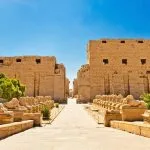 A wide, straight pathway lined with rows of Sphinx statues (the kebash Way), leading between the massive mud-brick pylons of the Karnak Temple under a bright blue sky.