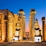 The illuminated Luxor Temple at Night, showing massive columns, statues of pharaohs, and the grand entrance courtyard bathed in warm yellow light against the dark blue sky.