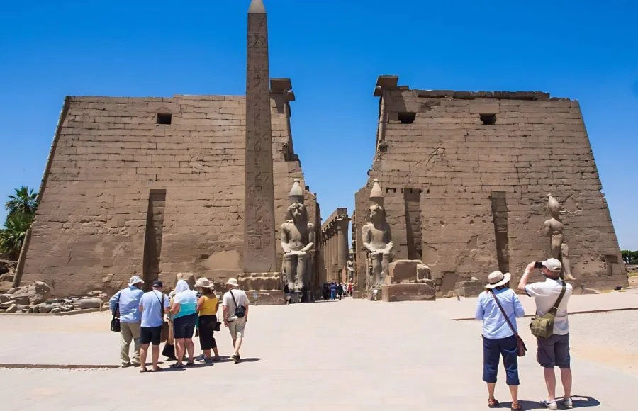 A group of Luxor Temple visitors stand in front of the massive first pylon, the standing statues of Ramesses II, and a tall obelisk under a clear blue sky.