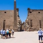 Tourists stand in front of the colossal statues and the massive facade of the Luxor Temple, admiring its grand entrance.