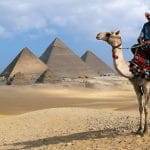 A Man Riding a Camel in traditional clothing is viewed from a low angle, silhouetted against the bright sky, with the Pyramids of Giza clearly visible in the desert background.