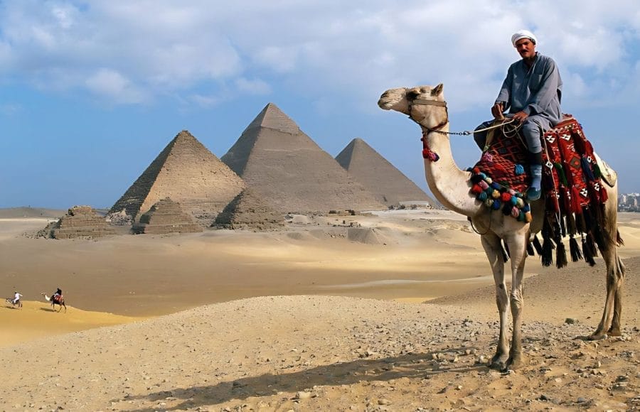 A Man Riding a Camel in traditional clothing is viewed from a low angle, silhouetted against the bright sky, with the Pyramids of Giza clearly visible in the desert background.