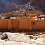 An external view of Saint Catherine Monastery Egypt, showing its high, fortified walls built into the base of a jagged, rocky mountain in the Sinai desert.