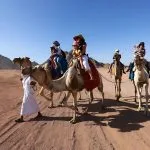 A group of tourists Riding Camels in Desert with a local guide in Sharm El-sheikh Egypt, showcasing the vast, flat desert landscape and distant rocky mountains.