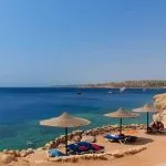 A beautiful view of Sharm el-Sheikh Beach, showing straw sun umbrellas and lounge chairs on a rocky beach overlooking the deep blue waters of the Red Sea.