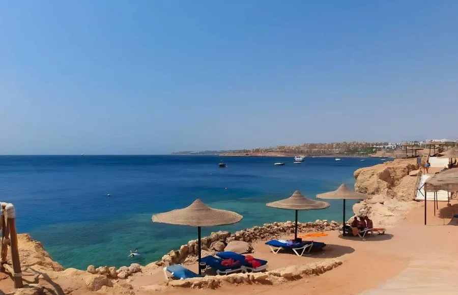 A beautiful view of Sharm el-Sheikh Beach, showing straw sun umbrellas and lounge chairs on a rocky beach overlooking the deep blue waters of the Red Sea.