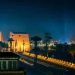 A nighttime photo of the Sound and Light Show Luxor, showing the illuminated Pylon and the Avenue of Sphinxes at Luxor Temple, glowing warmly against the dark blue sky.