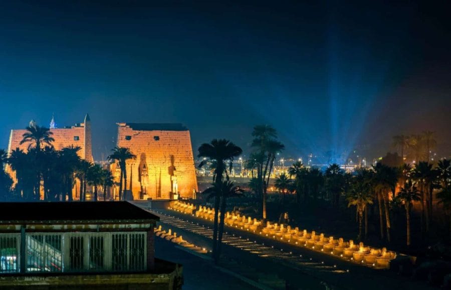 A nighttime photo of the Sound and Light Show Luxor, showing the illuminated Pylon and the Avenue of Sphinxes at Luxor Temple, glowing warmly against the dark blue sky.