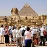 A group of Tourists stands near the smaller wall enclosures, looking toward the Sphinx and the massive Great Giza Pyramids in the background under a blue sky.