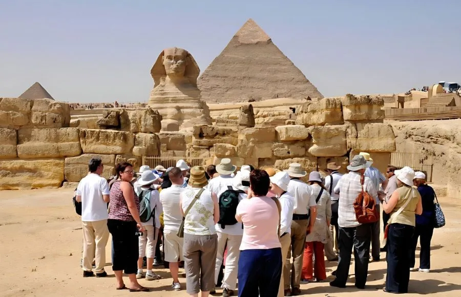 A group of Tourists stands near the smaller wall enclosures, looking toward the Sphinx and the massive Great Giza Pyramids in the background under a blue sky.