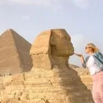 A Girl poses in the desert with her hand near her lips, appearing to Kiss the small Sphinx statue at Giza, with a large pyramid in the background.
