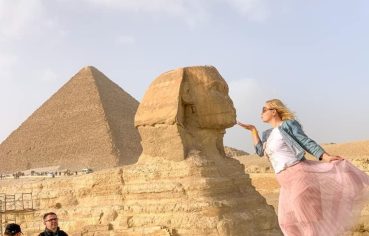 A Girl poses in the desert with her hand near her lips, appearing to Kiss the small Sphinx statue at Giza, with a large pyramid in the background.