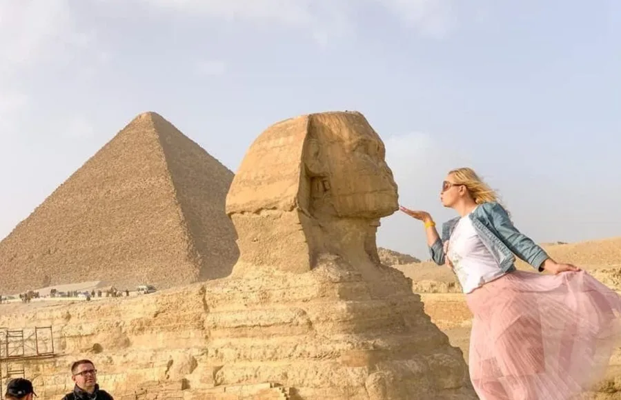 A Girl poses in the desert with her hand near her lips, appearing to Kiss the small Sphinx statue at Giza, with a large pyramid in the background.