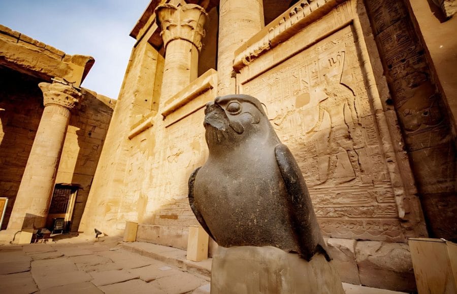 A massive, grey granite Statue of Horus Falcon God at Temple of Horus or Edfu Temple, standing guard in the courtyard with the temple's colossal pylon columns and reliefs in the background.