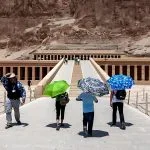 Tourists with umbrellas walking up the ramp on a Sunny Day at Temple Of Hatshepsut, a massive multi-terraced structure carved into the high desert cliff.