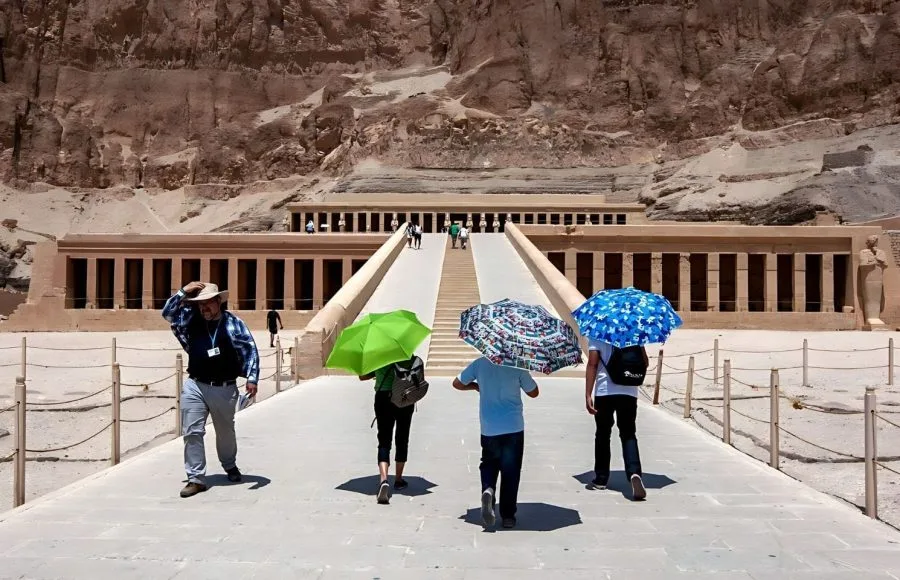 Tourists with umbrellas walking up the ramp on a Sunny Day at Temple Of Hatshepsut, a massive multi-terraced structure carved into the high desert cliff.