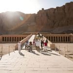 Tourists Taking Photos On Stairs Temple Of Hatshebsut, ascending the ramps towards the terraced facade of the magnificent mortuary temple, with sunlit cliffs in the background.