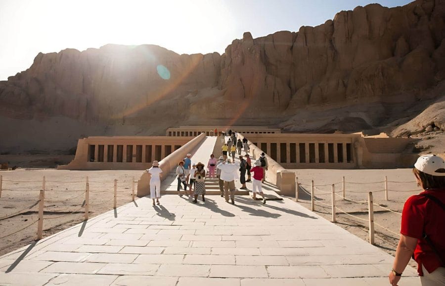 Tourists Taking Photos On Stairs Temple Of Hatshebsut, ascending the ramps towards the terraced facade of the magnificent mortuary temple, with sunlit cliffs in the background.