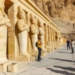 Two tourists are Taking Photos Temple Of Hatshepsut, standing among the colossal stone statues of the queen, with the high desert cliffs in the background.