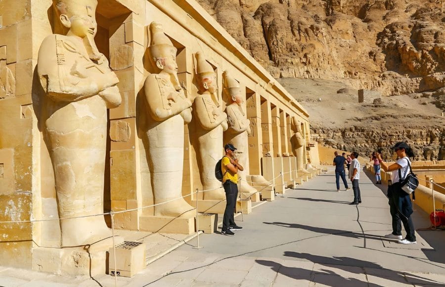 Two tourists are Taking Photos Temple Of Hatshepsut, standing among the colossal stone statues of the queen, with the high desert cliffs in the background.