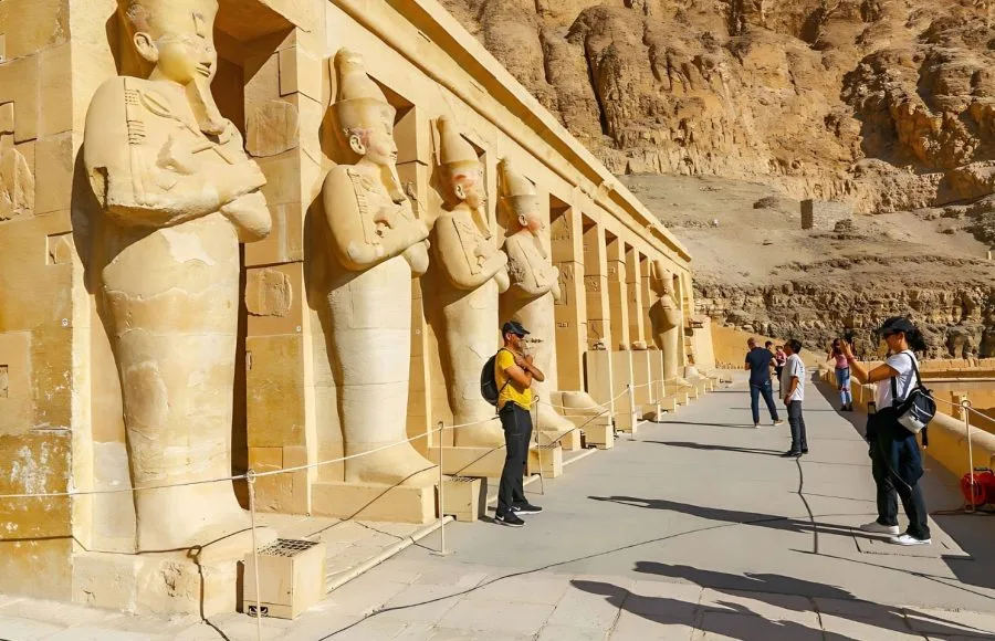 Two tourists are Taking Photos Temple Of Hatshepsut, standing among the colossal stone statues of the queen, with the high desert cliffs in the background.
